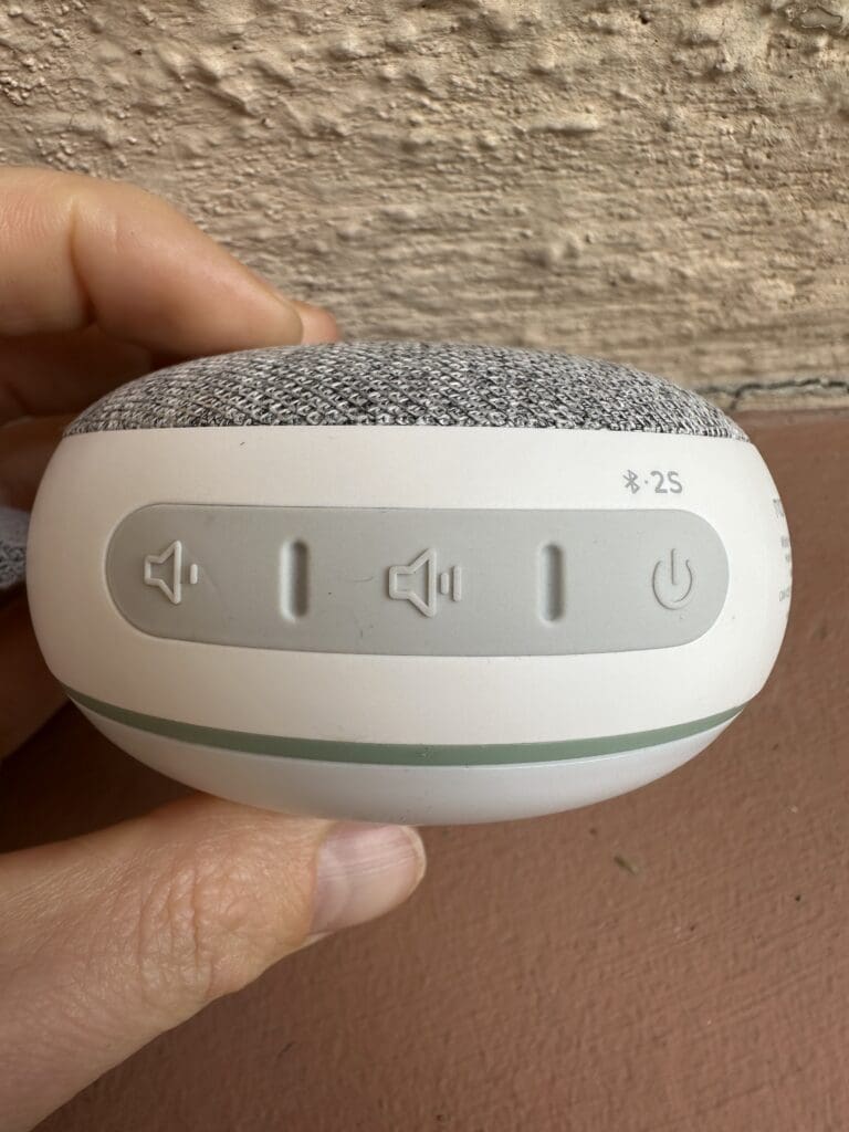 Momcozy portable baby sound machine hand holding it showing up close view of buttons (power, bluetooth, and volume)