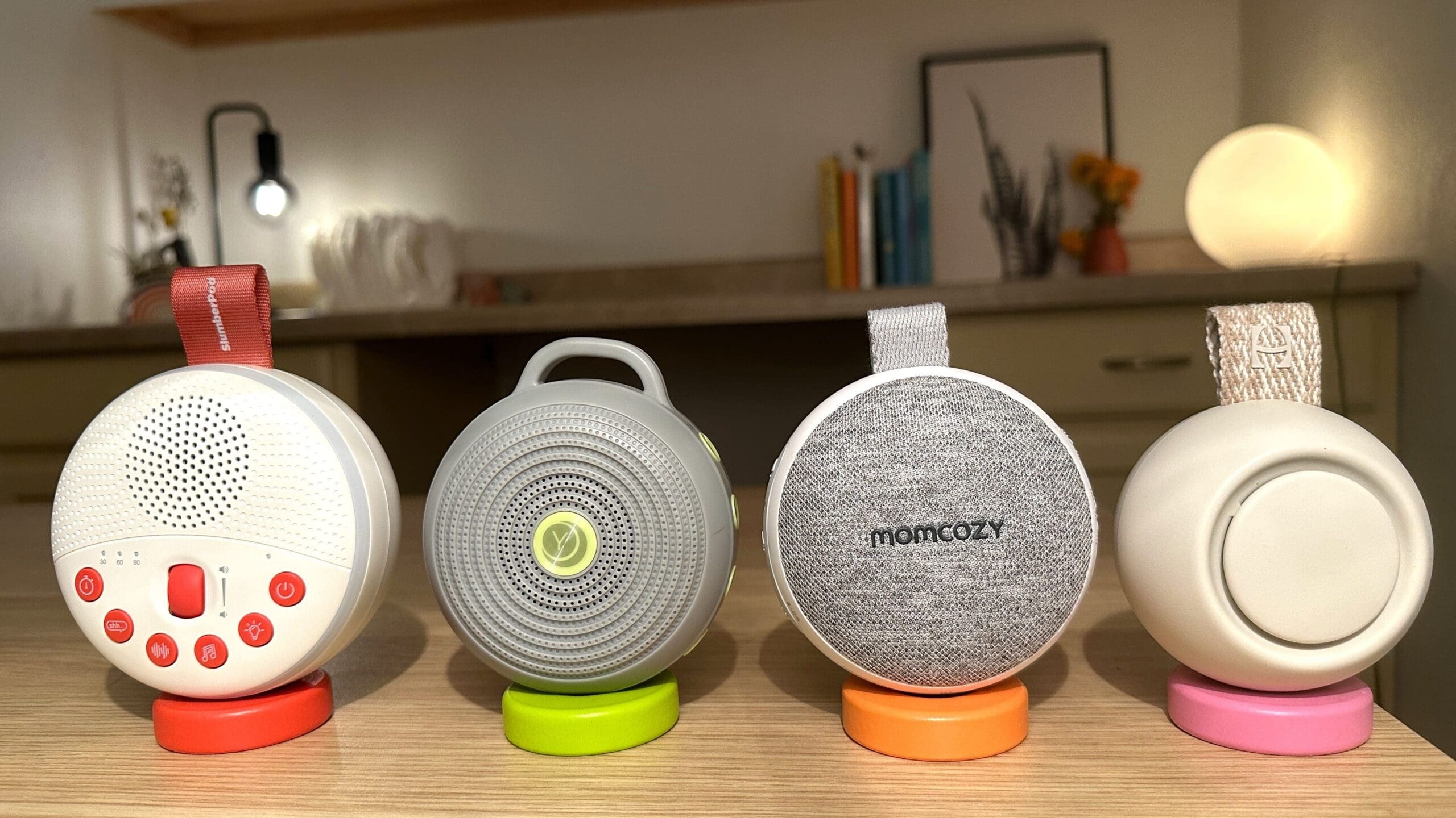 Front view of four popular portable baby sound machines up close front sitting on desk (SlumberPod SlumberCalm vs. Yogasleep Hushh vs. Momcozy Portable vs. Hatch Go)